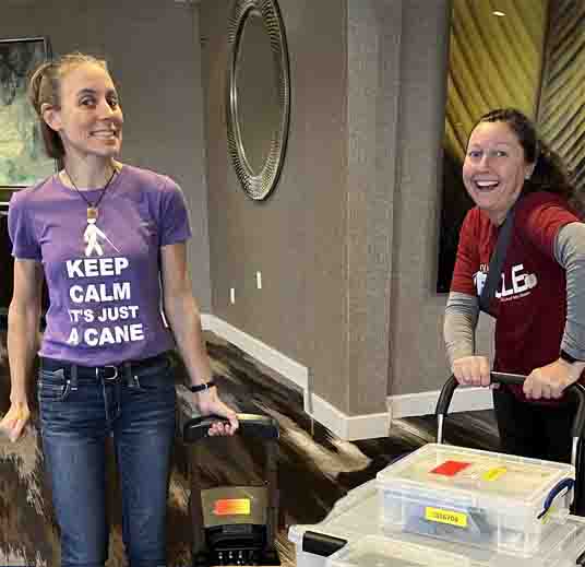 Sue and Andrea are laughing and pushing or pulling carts full of boxes and equipment.